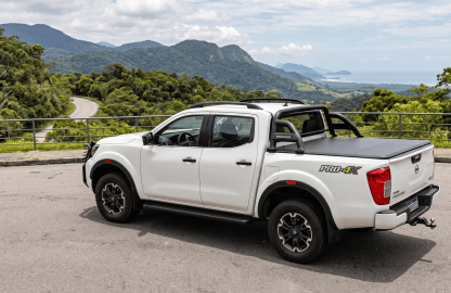 Uma picape Nissan Frontier branca equipada com capota marítima, santo antônio preto, estribo lateral e engate de reboque, estacionada em um mirante de estrada com vista para montanhas e o mar ao fundo.