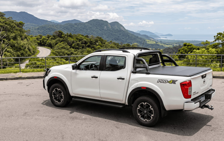 Uma picape Nissan Frontier branca equipada com capota marítima, santo antônio preto, estribo lateral e engate de reboque, estacionada em um mirante de estrada com vista para montanhas e o mar ao fundo.